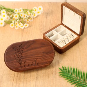 May include: A wooden jewelry box with an open lid displaying rings and a closed oval box with a floral design and the name "Emillh" engraved on it. The jewelry box is made of dark wood and has a white interior.