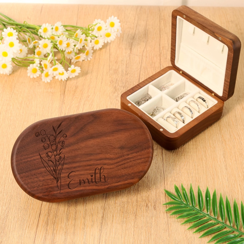 May include: A wooden jewelry box with an open lid displaying rings and a closed oval box with a floral design and the name "Emillh" engraved on it. The jewelry box is made of dark wood and has a white interior.