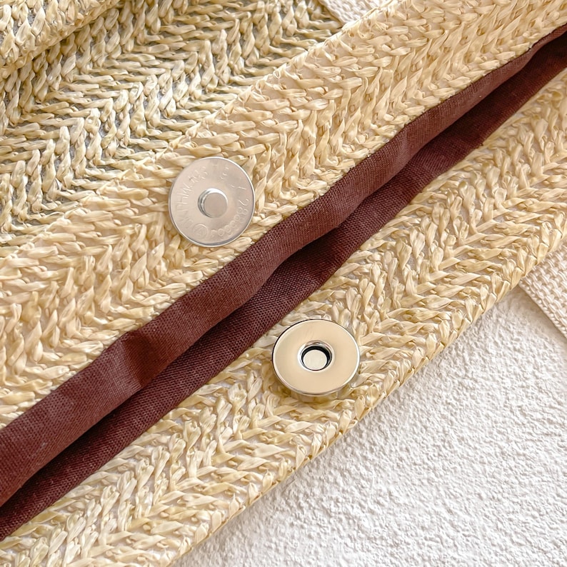 May include: Close-up of a woven straw bag with a herringbone pattern. The bag features a dark brown fabric lining and two silver magnetic closures. The straw has a natural, light beige color.