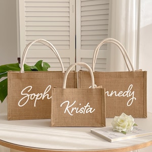 May include: Three burlap tote bags with white handles and personalized names in cursive script: "Soph", "Krista", and "nnedy". The bags are different sizes and sit on a white surface. A white rose and a small book are also on the surface.