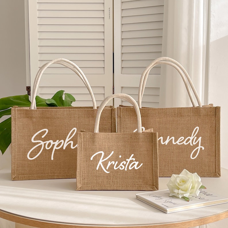 May include: Three burlap tote bags with white handles and personalized names in cursive script: "Soph", "Krista", and "nnedy". The bags are different sizes and sit on a white surface. A white rose and a small book are also on the surface.