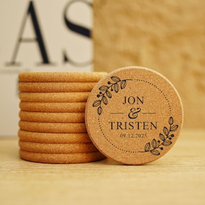 Puede incluir: Una pila de posavasos de corcho con un solo posavasos que presenta un diseño en blanco y negro de una corona de hojas y el texto "JON & TRISTEN 09.12.2025".
