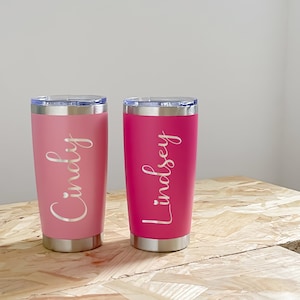 May include: Two stainless steel tumblers, one pink and one magenta, with silver lids. The pink tumbler has the name "Cindy" in a cursive font, and the magenta tumbler has the name "Lindsey" in a cursive font.