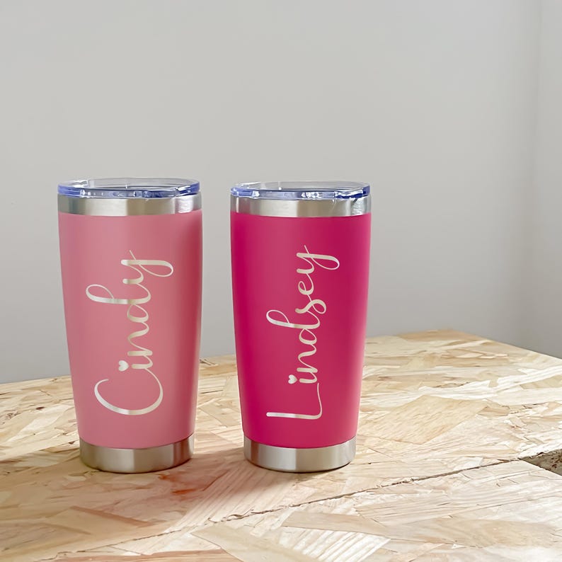 May include: Two stainless steel tumblers, one pink and one magenta, with silver lids. The pink tumbler has the name "Cindy" in a cursive font, and the magenta tumbler has the name "Lindsey" in a cursive font.
