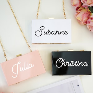 May include: Three small rectangular clutch purses with gold chains. The purses are white, black, and pink. Each purse has a name written in white script on the front: "Susanne", "Christina", and "Julia".