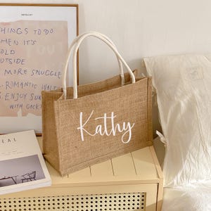May include: A rectangular, tan burlap tote bag with white handles. The name "Kathy" is written in white script across the front. The bag is sitting on a light-colored surface, with a framed print and a book visible in the background.
