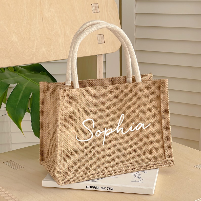 May include: A rectangular jute tote bag with white handles and the name "Sophia" in white script. The bag is sitting on a book with the words "Coffee or Tea" visible.