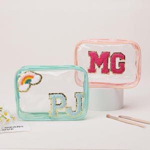May include: Two clear cosmetic bags. One is teal with a rainbow and the letters "PJ" in blue and gold. The other is pink with the letters "MG" in pink and gold. Both bags are rectangular and have a zipper closure.