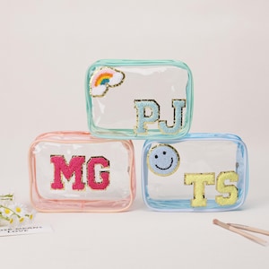 Op de afbeelding: Drie doorzichtige cosmetische tassen met kleurrijke randen en geborduurde patches. Eén tas heeft een regenboog, een andere de letters "PJ", en de derde de letters "MG". De laatste tas heeft een smiley en de letters "TS".