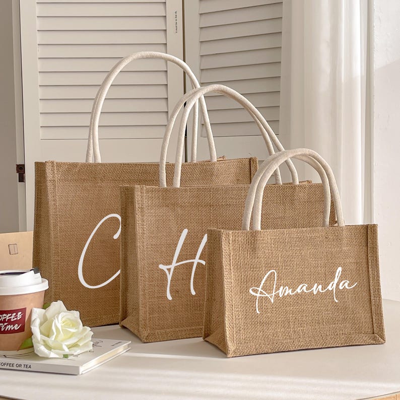 May include: Three burlap tote bags with white handles and lettering. The largest bag has the letter "C", the middle bag has "H", and the smallest bag has the name "Amanda" in a script font. A coffee cup and a white rose are in the foreground.