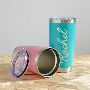 May include: Two stainless steel insulated tumblers, one pink and one teal. The teal tumbler has the name "Rachel" in a cursive font. Both tumblers have clear lids and silver rims. The tumblers are on a wooden surface.