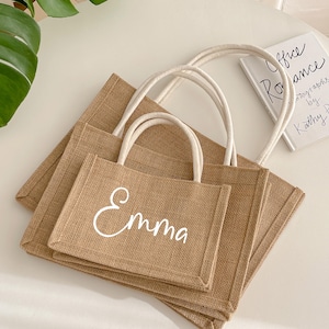 May include: Three rectangular burlap tote bags with white handles. The smallest bag has the name "Emma" in white script. A small book with the title "Office Romance" is visible in the background.