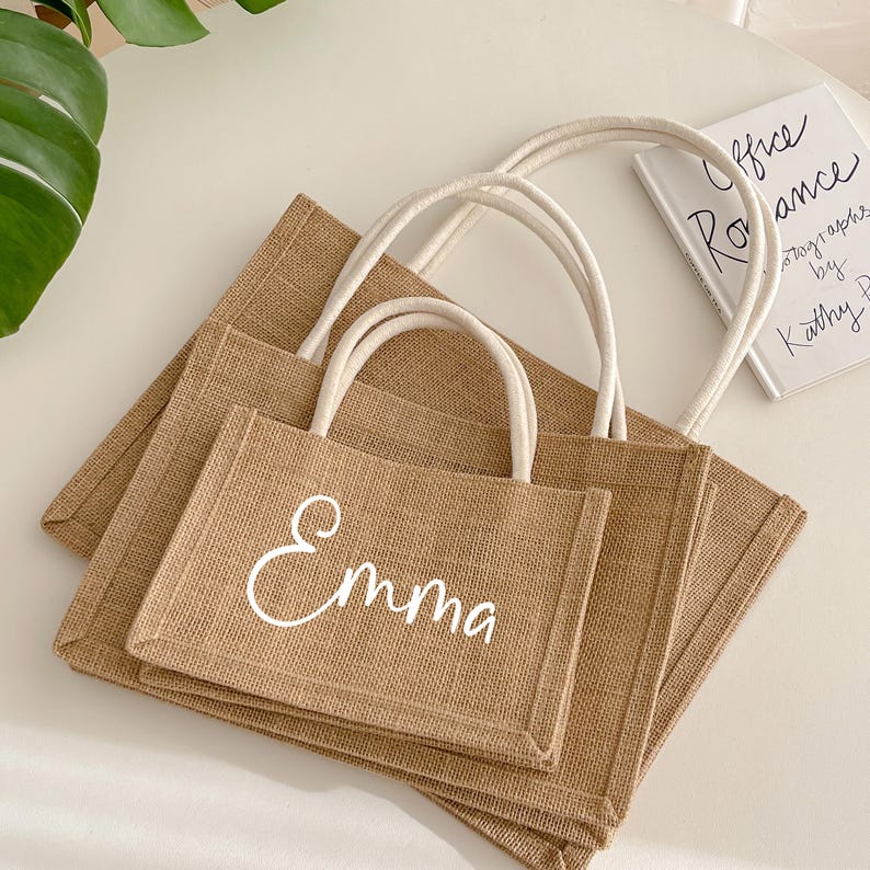 May include: Three rectangular burlap tote bags with white handles. The smallest bag has the name "Emma" in white script. A small book with the title "Office Romance" is visible in the background.