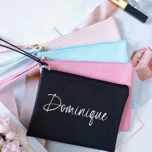 May include: Four zippered pouches in different colors, stacked on top of each other. The bottom pouch is black with the name "Dominique" written in white cursive lettering.