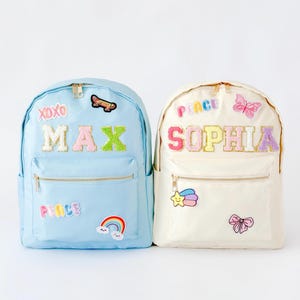 Puede incluir: Dos mochilas personalizadas. Una es azul claro con parches "MAX" y "PEACE", además de un arcoíris y una patineta. La otra es blanquecina con parches "SOPHIA" y "PEACE", una mariposa, una estrella y un lazo.