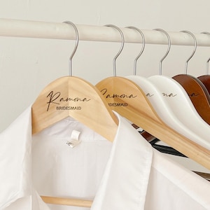 May include: A row of wooden clothes hangers with metal hooks, each engraved with a name and the word "BRIDESMAID". The hangers are in shades of wood, white, and brown, and are holding a white shirt.