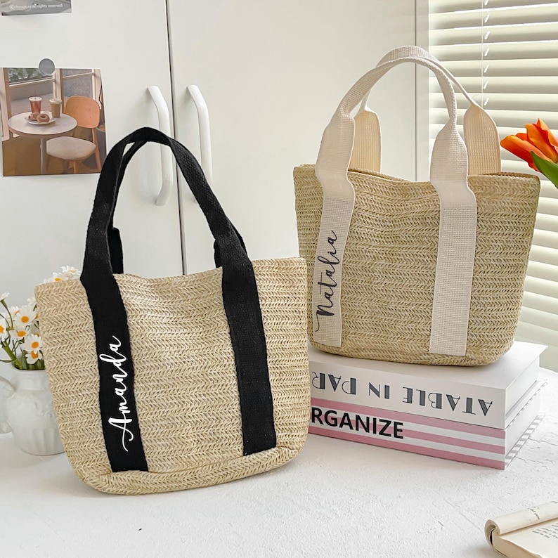 May include: Two woven straw tote bags. One has black handles and the name "Amanda" in white script. The other has cream-colored handles and the name "Natalia" in white script. Both bags are rectangular with a woven design.