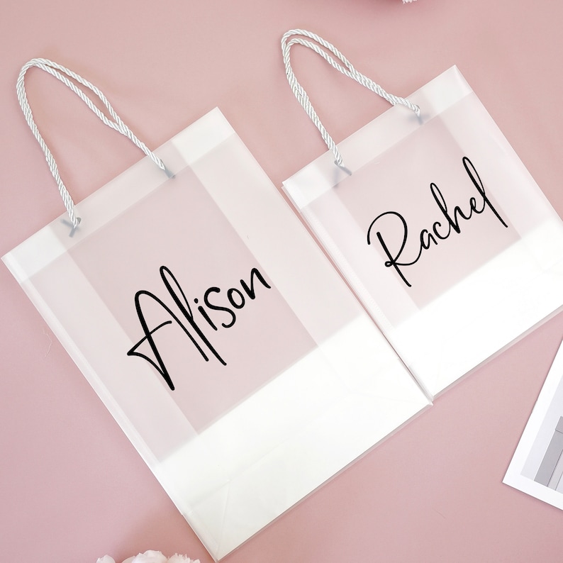 May include: Two clear plastic gift bags with white rope handles. The bags have white borders and the names "Alison" and "Rachel" written in black cursive.