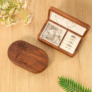 May include: A wooden jewelry box with an open lid, revealing necklaces and rings. The box is brown with a white interior. The closed lid has a floral design and the name "Rose" engraved on it. The jewelry box is on a wooden surface.
