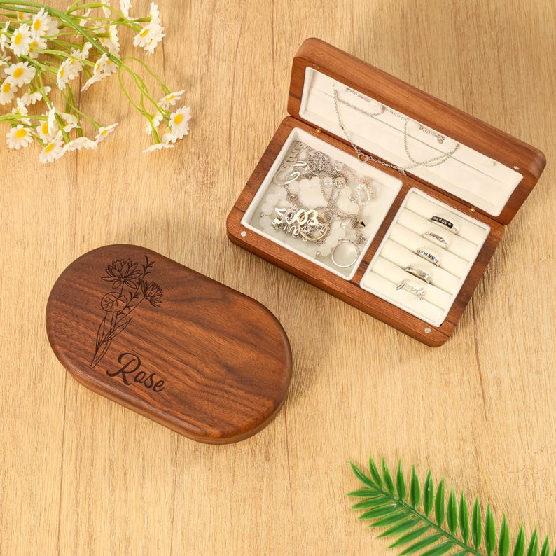 May include: A wooden jewelry box with an open lid, revealing necklaces and rings. The box is brown with a white interior. The closed lid has a floral design and the name "Rose" engraved on it. The jewelry box is on a wooden surface.