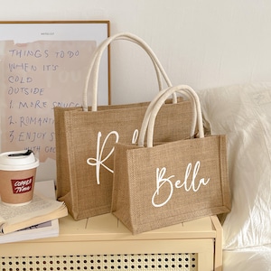 May include: Two burlap tote bags with white handles. The larger bag has the letter "K" in white script, and the smaller bag says "Bella". The bags are sitting on a light-colored surface.