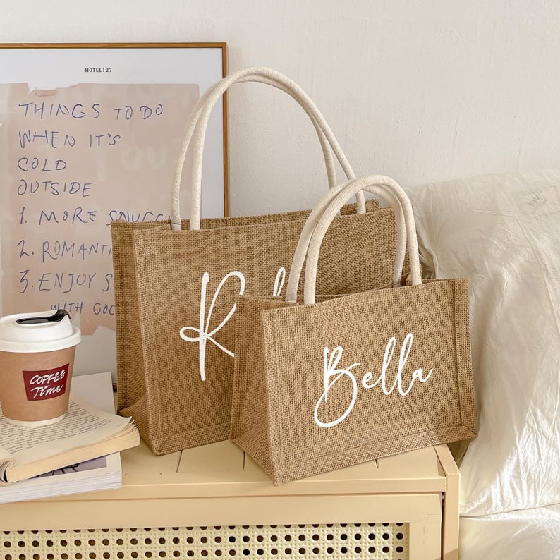 May include: Two burlap tote bags with white handles. The larger bag has the letter "K" in white script, and the smaller bag says "Bella". The bags are sitting on a light-colored surface.