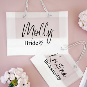 May include: Two white gift bags with clear plastic windows and white handles. The top bag has the text "Molly Bride" and the bottom bag has the text "Kristina Bridesmaid".