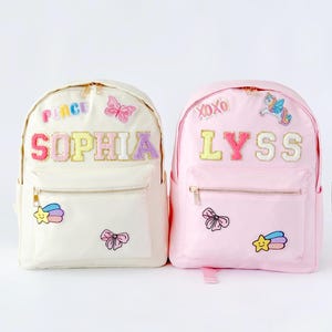 Könnte beinhalten: Zwei Kinderrucksäcke, einer in Creme und einer in Rosa. Der cremefarbene Rucksack hat den Namen "SOPHIA" und einen Schmetterlingsaufnäher. Der rosa Rucksack hat den Namen "LYSS" und einen Einhornaufnäher. Beide haben goldene Reißverschlüsse und Vordertaschen.