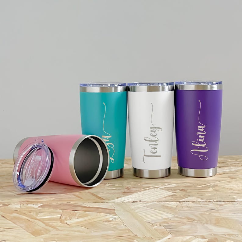 May include: A collection of personalized tumblers in various colors: pink, teal, white, and purple. Each tumbler has a silver rim and lid, with a name elegantly scripted on the front. The tumblers are displayed on a wooden surface.