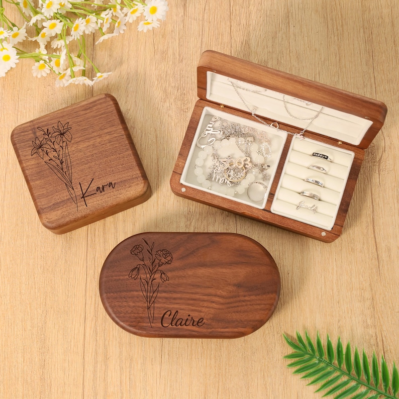 May include: Wooden jewelry boxes, one open, displaying necklaces and rings. Two closed boxes feature floral designs and names: "Kara" and "Claire." The boxes are a warm brown color, with a light-colored interior in the open box.