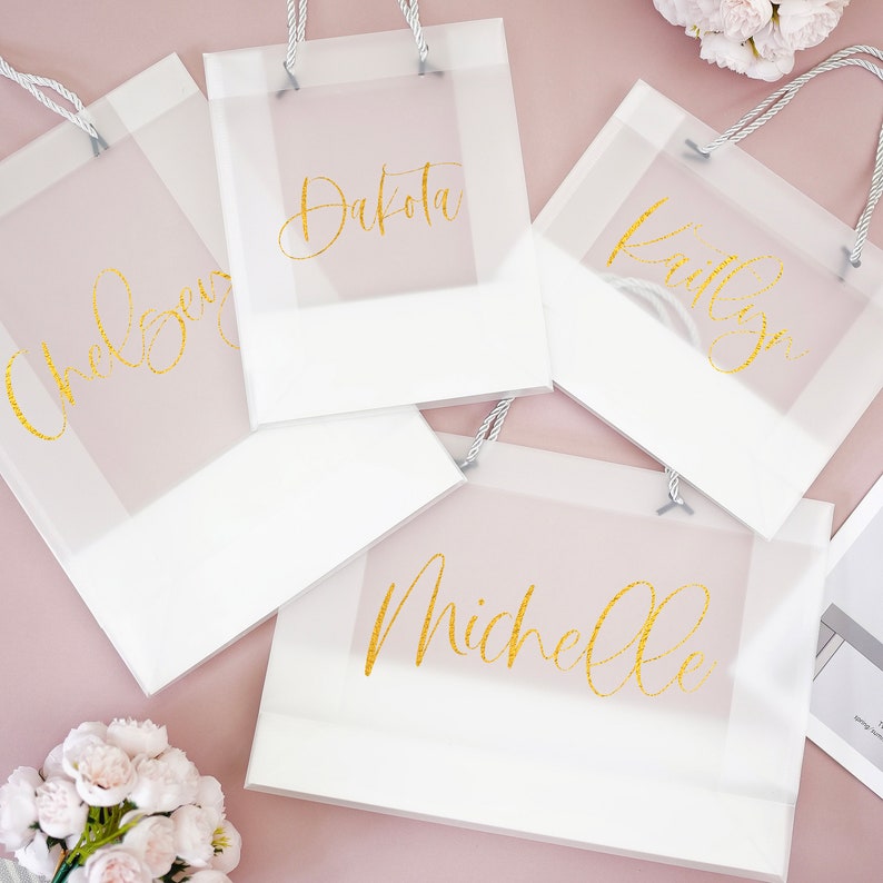 May include: Four white gift bags with clear cellophane windows and gold lettering. The names Dakota, Bailey, Michelle, and Chelsea are written on the bags.