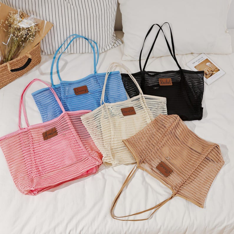 Puede incluir: Una colecci&oacute;n de bolsos tote de malla en varios colores: rosa, azul, crema, negro y marr&oacute;n claro. Cada bolso tiene una etiqueta rectangular de cuero con un nombre. Los bolsos tienen asas largas y se muestran sobre una superficie blanca.
