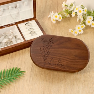 May include: A wooden jewelry box with an open lid, revealing compartments for rings and necklaces. The lid of the box features an engraved floral design and the name "Andrea". The box is made of dark brown wood.