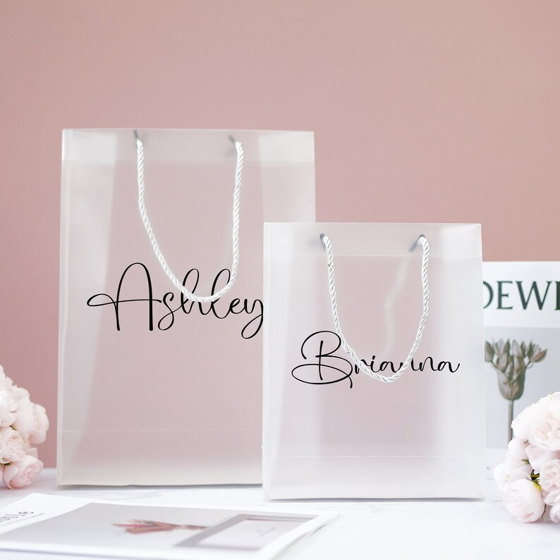 May include: Two clear plastic gift bags with white rope handles. The larger bag has the name "Ashley" printed on it in black script. The smaller bag has the name "Briana" printed on it in black script.