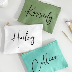 Può includere: Tre trousse cosmetiche personalizzate in tela verde, bianca e turchese. Ogni borsa ha una cerniera dorata e un nome stampato in corsivo nero: Kassidy, Hailey e Colleen. Pennelli per il trucco sono visibili sullo sfondo.