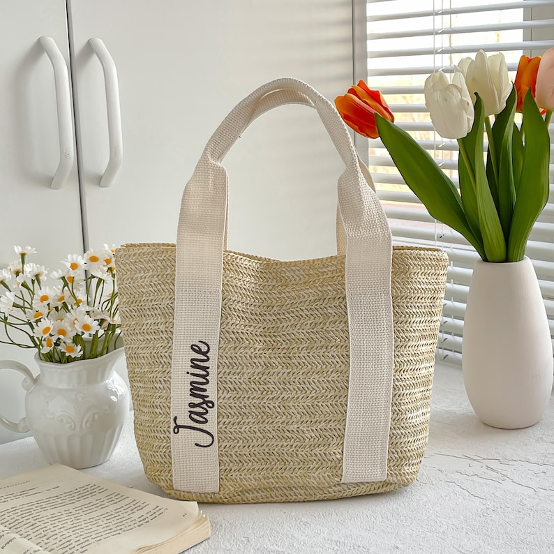 May include: A woven straw tote bag with cream-colored handles and the name "Jasmine" printed vertically on the side. The bag is rectangular with a wide opening. A vase of flowers is in the background.