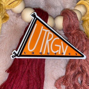 May include: An orange sticker with the text "UTRGV Vaqueros" in white, inside a black-outlined triangle. The sticker is set against a backdrop of colourful tassels and wooden beads, with shades of yellow, burgundy, and pink.