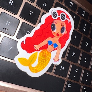 May include: A sticker of a cartoon mermaid with red hair, sunglasses, and a yellow tail. The mermaid is wearing a blue seashell top and has a heart tattoo. The sticker is on a laptop keyboard.