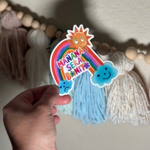 May include: A vibrant sticker with a rainbow, sun, and clouds, displaying the text "MAÑANA SERÁ BONITO." The rainbow is multi-coloured, and the sun has a smiling face. The sticker includes a blue cloud with a sad face and a happy cloud.