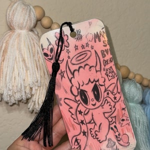 May include: A pink and white bookmark with a black and white illustration of a cartoon angel with horns. The bookmark has a black tassel and the text "Made By Chicaho "EEZ" on it.