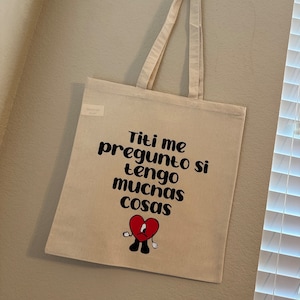 Op de afbeelding: Een beige canvas draagtas met twee schouderbanden. De voorzijde is voorzien van zwarte tekst met de tekst Titi me pregunto si tengo muchas cosas en een afbeelding van een verdrietig rood hart met zwarte armen, benen en een wit verband op het gezicht.