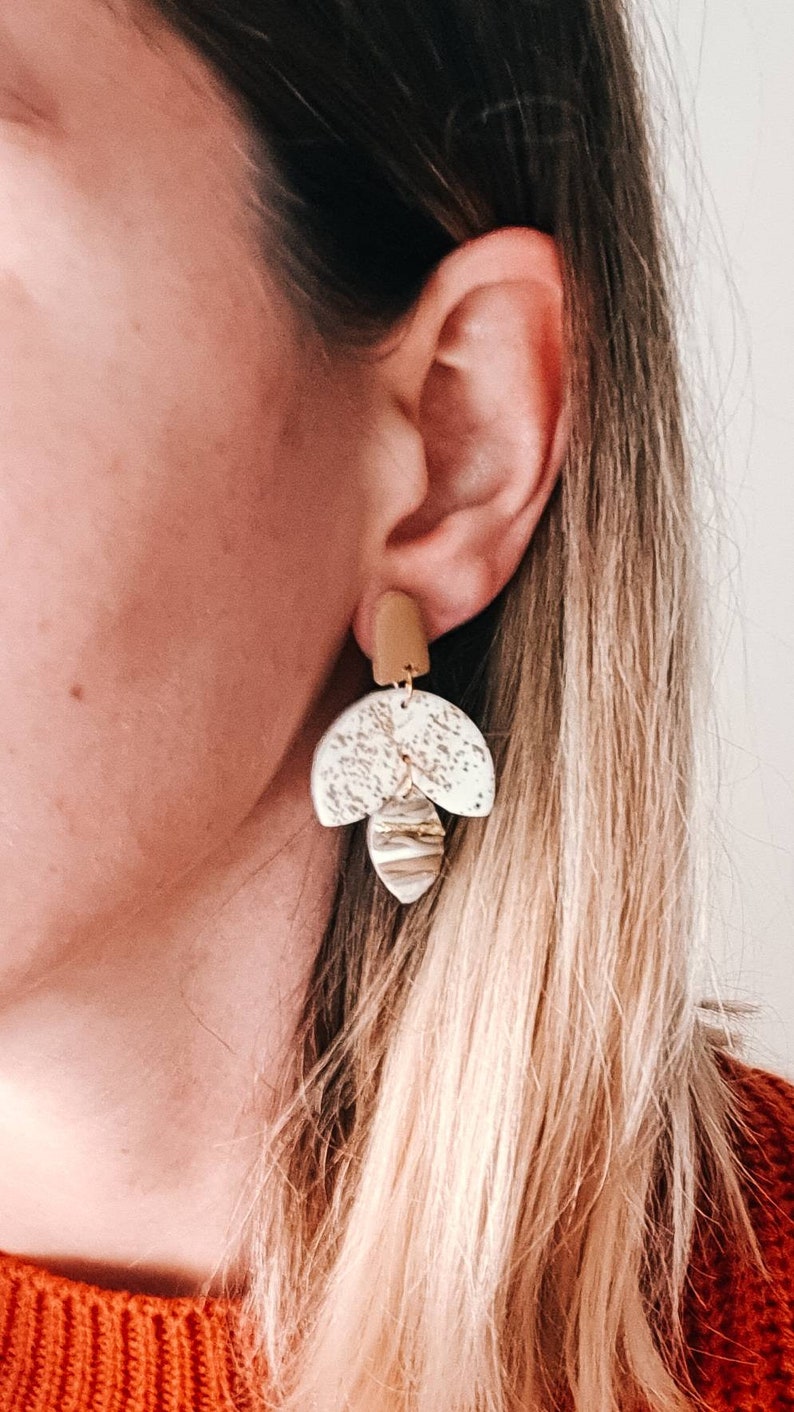 Op de afbeelding: Een paar oorhangers met een geometrisch ontwerp in wit en goud. De oorhangers zijn gemaakt van klei en hebben een textuur.
