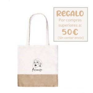 Op de afbeelding: Een witte tote bag met een bruine jute bodem. De tas heeft een zwarte lijntekening van een vrouwen gezicht met de tekst "fuinua clay" eronder. De tas is een cadeau voor aankopen boven de 50 euro. De tekst "REGALO Por compras superiores a: 50&euro; (Sin contar envio)" is in perzikkleur.