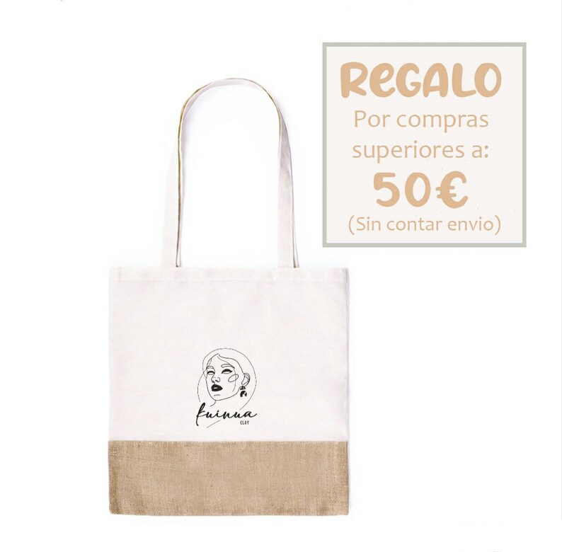 Op de afbeelding: Een witte tote bag met een bruine jute bodem. De tas heeft een zwarte lijntekening van een vrouwen gezicht met de tekst "fuinua clay" eronder. De tas is een cadeau voor aankopen boven de 50 euro. De tekst "REGALO Por compras superiores a: 50&euro; (Sin contar envio)" is in perzikkleur.