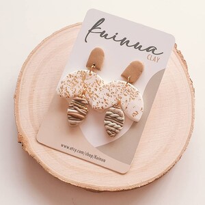 Op de afbeelding: Een paar gouden en witte kleioorbellen met een bruin en gouden gestreept ontwerp. De oorbellen bevinden zich op een witte kaart met de tekst "Kiuina Clay" en "www.etsy.com/shop/Kiuina".