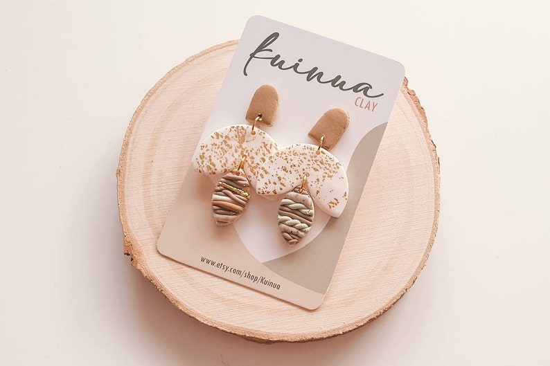 Op de afbeelding: Een paar gouden en witte kleioorbellen met een bruin en gouden gestreept ontwerp. De oorbellen bevinden zich op een witte kaart met de tekst "Kiuina Clay" en "www.etsy.com/shop/Kiuina".