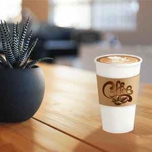 Pu&ograve; includere: Una tazza di caff&egrave; di carta bianca con una manica di cartone marrone con la scritta "Coffee" e chicchi di caff&egrave;. La tazza contiene un latte con un design decorativo in schiuma. Una pianta succulenta in vaso &egrave; su un tavolo di legno.