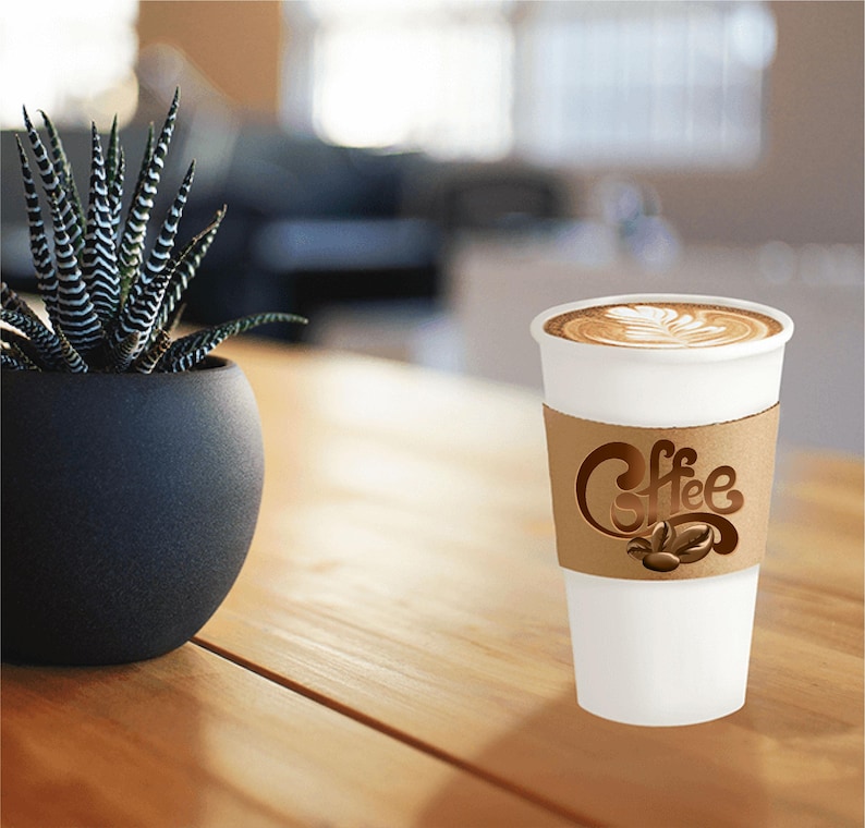 Pu&ograve; includere: Una tazza di caff&egrave; di carta bianca con una manica di cartone marrone con la scritta "Coffee" e chicchi di caff&egrave;. La tazza contiene un latte con un design decorativo in schiuma. Una pianta succulenta in vaso &egrave; su un tavolo di legno.