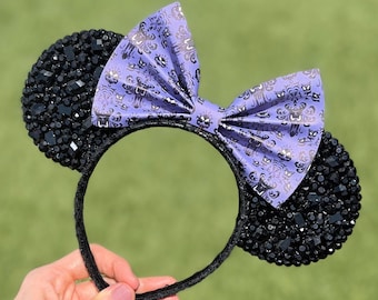 Op de afbeelding: Paarse en zwarte Minnie Mouse oren met een Haunted Mansion-thema strik. De strik is bedekt met een paarse en zilveren print met een herhalend patroon van geesten.