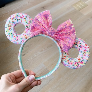 May include: A pair of pink and white sprinkle donut-themed Minnie Mouse ears with a pink sequin bow.